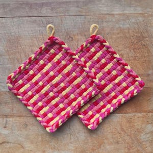 May include: Two woven potholders in shades of pink, red, and yellow. Each square-shaped potholder has a braided edge and a loop for hanging. The potholders are displayed on a wooden surface.