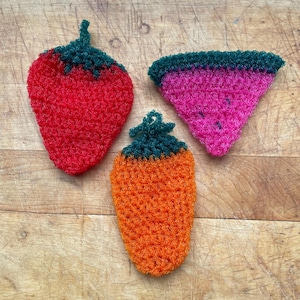 May include: Three crocheted fruit-shaped items: a red strawberry, an orange carrot, and a pink watermelon slice. Each has a green top. The items are arranged on a wooden surface, showcasing handmade crafts.
