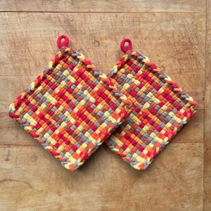 May include: Two square woven potholders in a vibrant pattern of red, orange, yellow, and gray. Each potholder has a small red loop for hanging. The potholders are made of a thick, textured material.