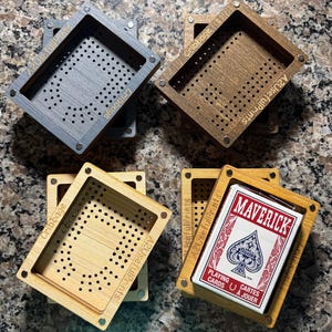 May include: Four rectangular wooden cribbage boards with magnetic corners. Three boards are empty, showing a grid of holes and the text "Cribbage" and "A2UsefulPrints". One board contains a deck of Maverick playing cards with red and blue graphics.