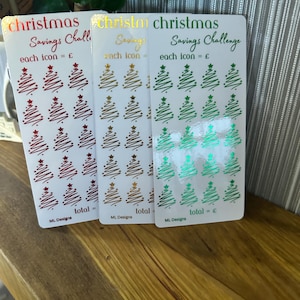 Puede incluir: Tres hojas de desafío de ahorro navideño con un total de 24 iconos de árboles de Navidad. Cada hoja tiene un tema de color diferente: rojo, dorado y verde. El texto en las hojas dice "Christmas Savings Challenge" y "each icon = £" o "each icon = €".