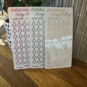 Puede incluir: Tres tarjetas de desafío de ahorro navideño con fondo blanco e iconos de árboles de Navidad dorados, verdes y rojos. El texto en las tarjetas dice "Desafío de ahorro navideño" y "cada icono = £".