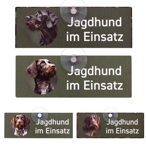 Könnte beinhalten: Rechteckige Schilder mit einem Hundeporträt und dem Text "Jagdhund im Einsatz" in Weiß. Die Schilder haben einen dunkelgrünen Hintergrund und sind mit Saugnäpfen befestigt. Die Hundeporträts variieren in Farbe und Rasse.