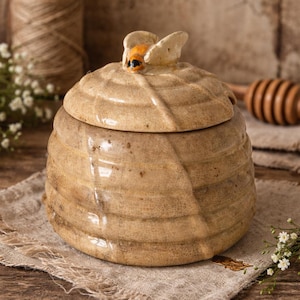Könnte beinhalten: Ein beigefarbener Honigtopf aus Keramik mit gerippter, an einen Bienenstock erinnernder Textur und passendem Deckel. Der Deckel ist mit einer kleinen, geformten Biene mit orangefarbenen und schwarzen Details verziert. Der Topf hat eine glänzende Oberfläche.