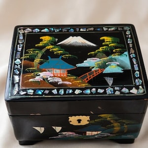 May include: A black rectangular lacquer jewelry box featuring a lid with a painted landscape of Mount Fuji, a red bridge, and a building, framed by inlaid iridescent mother-of-pearl pieces. The front panel displays a matching painted scene.