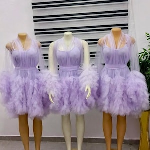 May include: Three lilac dresses displayed on mannequins. Each dress has a layered, ruffled skirt, a fitted bodice, and sheer sleeves. The dresses are set against a neutral backdrop, highlighting their design and texture.