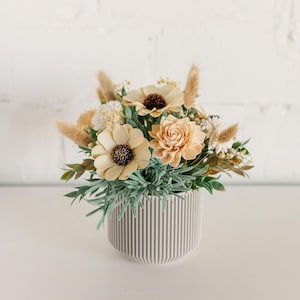 May include: A small, gray, striped ceramic planter with a floral arrangement of beige and brown flowers. The flowers are made of wood and have a rustic, natural look.