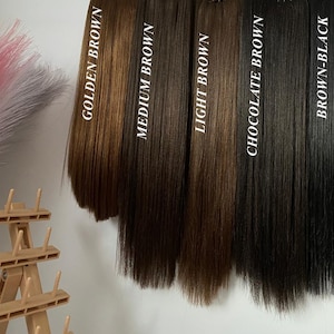 Op de afbeelding: Zes verschillende tinten haarverlengingen, hangend aan een rek. De kleuren zijn Goudbruin, Medium Bruin, Lichtbruin, Chocoladebruin, Bruin-Zwart en Zwart.