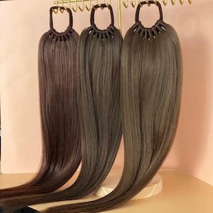May include: Three brown hair extensions with elastic bands. The hair extensions are hanging from a rack and are arranged in a row.