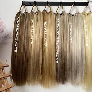 Könnte beinhalten: Sechs verschiedene Blondtöne von Haarverlängerungen, die an einem schwarzen Gestell hängen. Die Farbtöne sind mit "Bronde Highlights", "Honey Blonde", "Golden Blonde", "Blonde Dark Highlights", "Ash Blonde" und "Bleach Blonde" beschriftet.