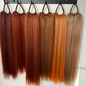 May include: Seven different shades of red hair extensions, hanging from a black rack. The shades range from dark auburn to light copper.