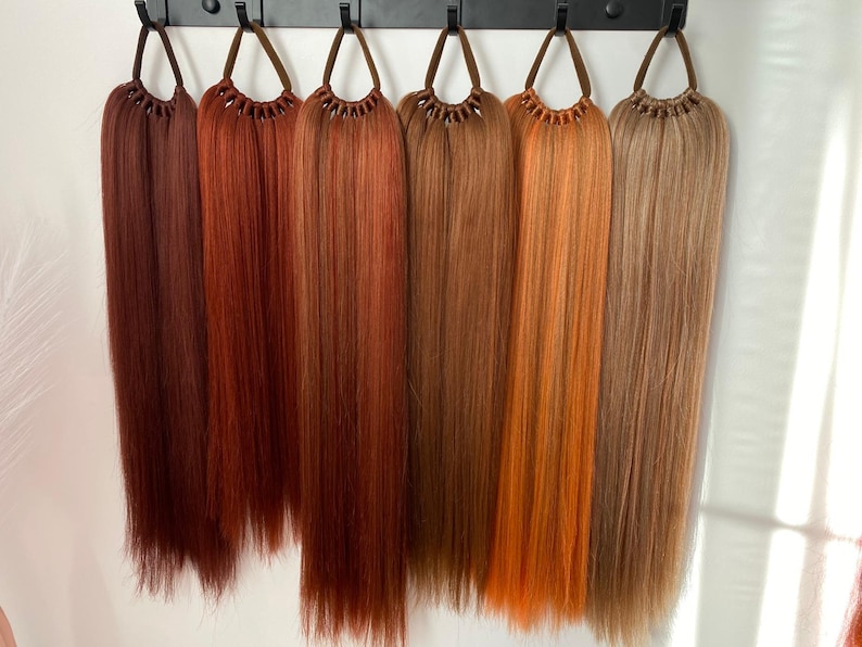 May include: Seven different shades of red hair extensions, hanging from a black rack. The shades range from dark auburn to light copper.