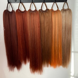 May include: Seven different shades of brown and red hair extensions hanging from hooks. The extensions are long and straight.