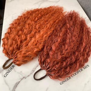 Può includere: Due ciocche di extension per capelli ricci nelle tonalità arancione e rame. La ciocca di sinistra è etichettata "GINGER" e quella di destra "GINGER COOPER MIX". Ogni ciocca è fissata con un elastico marrone.