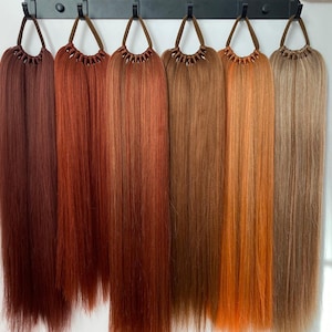 May include: Seven different shades of brown hair extensions hanging from hooks. The shades range from dark brown to light blonde.