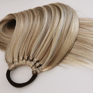 May include: A long, thick ponytail of blonde hair with light brown highlights, tied with a black elastic hair tie.