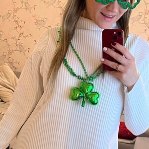 May include: A white ribbed sweater with a green shamrock necklace and green shamrock glasses. The necklace has the words "Happy St. Patrick's Day" on it.