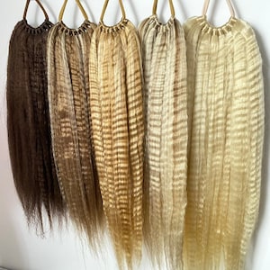 Op de afbeelding: Vijf verschillende tinten bruin, blond en lichtblond echt haar extensions hangend aan haken. De haarextensions zijn golvend en hebben een natuurlijke textuur.