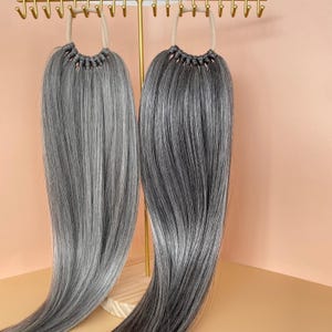 May include: Two bundles of gray and silver hair extensions hanging from a gold-colored display stand. The hair is straight and has a natural, blended appearance. The extensions are designed to add length and volume to hair.