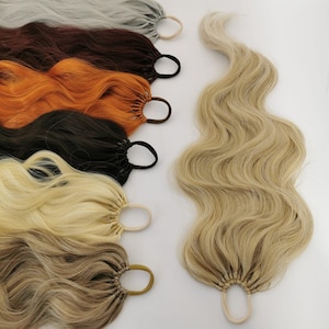 Könnte beinhalten: Eine Auswahl an welligen Haarverlängerungen in verschiedenen Farben, darunter Grau, Braun, Orange, Schwarz, Blond und Hellbraun. Jede Verlängerung ist mit einem Haargummi befestigt. Sie sind so konzipiert, dass sie Frisuren mehr Länge und Volumen verleihen.