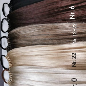 May include: Four different shades of human hair extensions, from black to blonde, with elastic bands at the top. The hair extensions are labeled with numbers: Nr. 0, Nr. 22, Nr. 12+22, Nr. 6, and Nr. 1.