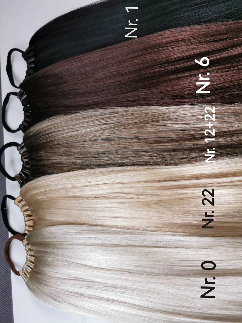 May include: Four different shades of human hair extensions, from black to blonde, with elastic bands at the top. The hair extensions are labeled with numbers: Nr. 0, Nr. 22, Nr. 12+22, Nr. 6, and Nr. 1.