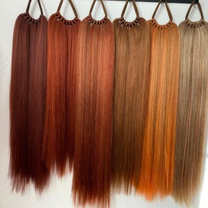 May include: Seven different shades of brown hair extensions, each with a brown elastic band at the top. The hair extensions are long and straight.