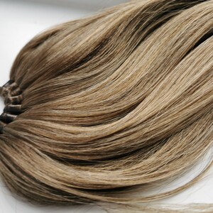 May include: A long, thick ponytail hair extension in a light brown shade with blonde highlights. The hair is tied with a black elastic band.