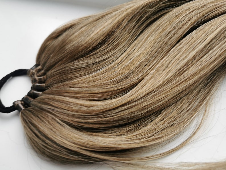 May include: A long, thick ponytail hair extension in a light brown shade with blonde highlights. The hair is tied with a black elastic band.