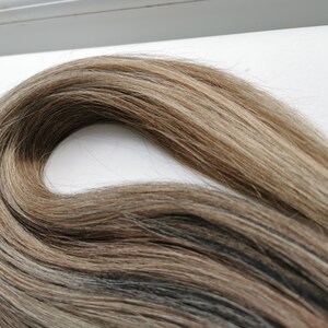 May include: A close-up of a bundle of light brown human hair extensions with blonde highlights. The hair is soft and silky, and it is arranged in a loose wave.