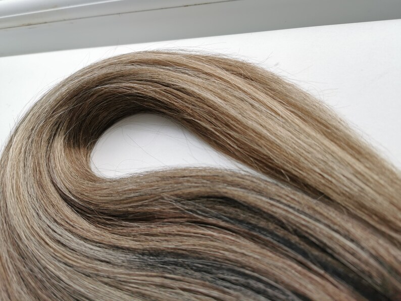 May include: A close-up of a bundle of light brown human hair extensions with blonde highlights. The hair is soft and silky, and it is arranged in a loose wave.