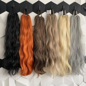 May include: Six different coloured hair extensions hanging on hooks. The extensions are wavy and range in colour from black to light blonde to grey.
