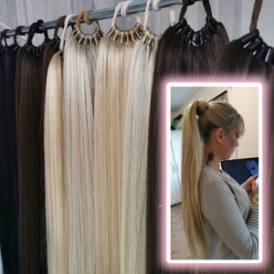 Op de afbeelding: Een selectie van haar extensions in verschillende tinten blond en bruin, hangend aan een rek. De extensions zijn lang en recht, en ze zijn allemaal met een zwart elastiekje aan elkaar gebonden. Een close-up van een vrouw die een blonde haar extension in een paardenstaart draagt.