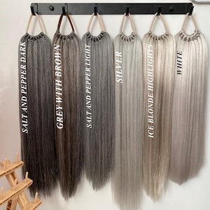 Puede incluir: Seis extensiones de cabello de diferentes tonos de gris y rubio, colgando de un estante negro. Los tonos están etiquetados como "Salt and Pepper Dark", "Grey with Brown", "Salt and Pepper Light", "Silver", "Ice Blonde Highlights" y "White".