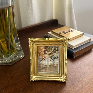 May include: A gold-framed picture with a cross-stitch design of a ballerina in a white dress. The ballerina is holding a bouquet of flowers and is surrounded by green foliage.