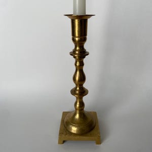 May include: A gold metal candlestick with a square base and a single candle holder.