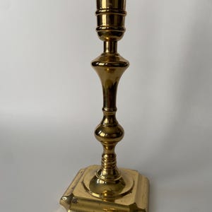 May include: A gold metal candlestick with a square base and a fluted design. The candlestick is tall and slender, with a simple, elegant design.