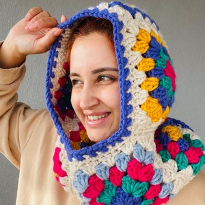 Puede incluir: Una capucha de ganchillo multicolor, con un dise&ntilde;o de cuadrados de la abuela. La capucha presenta una vibrante mezcla de colores, incluyendo azul, amarillo, rosa y verde, con una base crema y un borde azul. La capucha est&aacute; dise&ntilde;ada para ser usada.