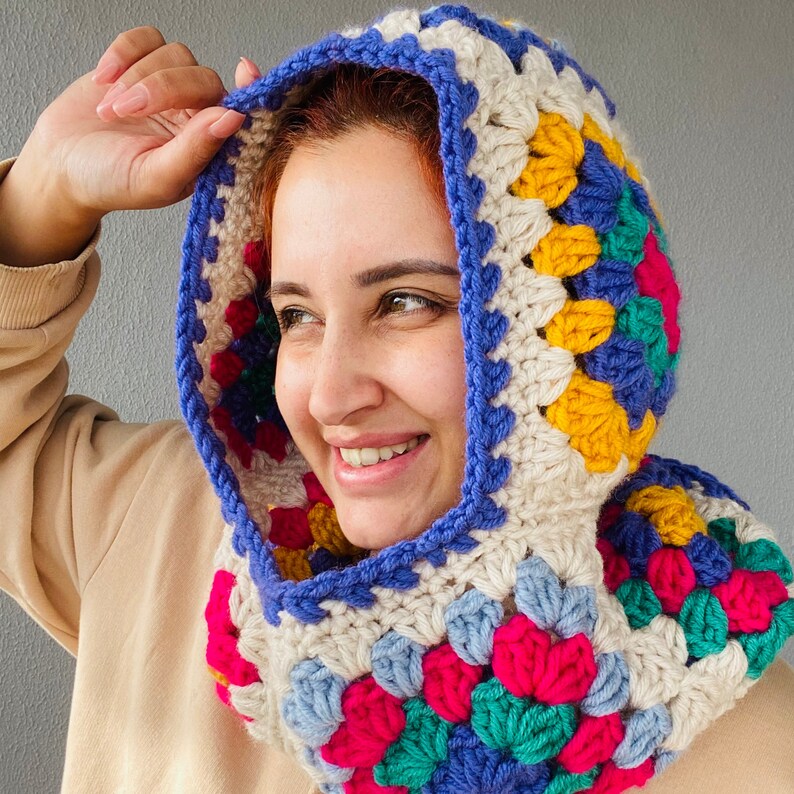 Puede incluir: Una capucha de ganchillo multicolor, con un dise&ntilde;o de cuadrados de la abuela. La capucha presenta una vibrante mezcla de colores, incluyendo azul, amarillo, rosa y verde, con una base crema y un borde azul. La capucha est&aacute; dise&ntilde;ada para ser usada.