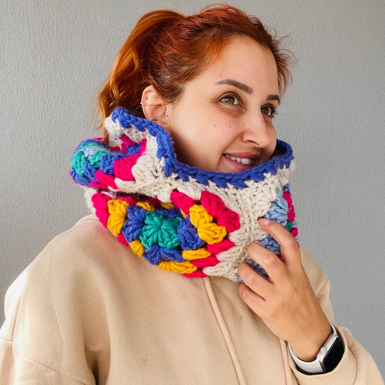 Puede incluir: Un cuello de ganchillo colorido con un patr&oacute;n de granny square. El cuello presenta una mezcla de colores brillantes, incluyendo rosa, azul, amarillo y verde, con una base crema y un borde azul. El calentador de cuello hecho a mano est&aacute; dise&ntilde;ado para ser usado alrededor del cuello.