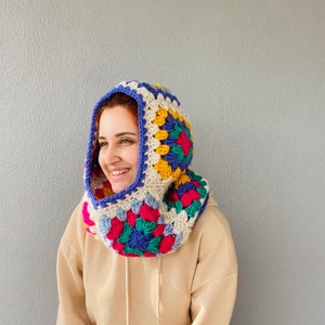 Puede incluir: Una colorida capucha de ganchillo con un patr&oacute;n de cuadrados de la abuela. La capucha presenta una vibrante mezcla de colores que incluyen azul, rosa, amarillo y crema, con un borde azul. La capucha hecha a mano est&aacute; dise&ntilde;ada para usarse alrededor del cuello y sobre la cabeza.