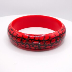 May include: A bright red bangle bracelet featuring a prominent black crackle pattern that covers the entire outer surface. The interior of the circular bracelet is a solid, smooth red finish, providing a contrast to the textured black design.