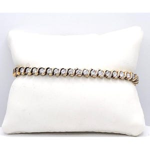 May include: A gold-tone tennis bracelet featuring a continuous row of round, clear, faceted stones set within an S-shaped metal link design. The bracelet is displayed horizontally across a plain, white, rectangular jewelry display pillow.
