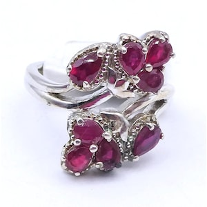 May include: A silver ring featuring two clusters of pear-shaped, deep red rubies. Each cluster contains three faceted stones set in metal prongs. The band of the ring has a smooth, polished finish and a bypass design that connects the two ruby floral motifs.