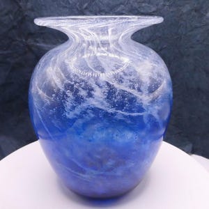 May include: A hand-blown glass vase featuring a rounded body and a wide, flared rim. The glass displays a deep blue base color with swirling white patterns and numerous small air bubbles embedded throughout the translucent material.