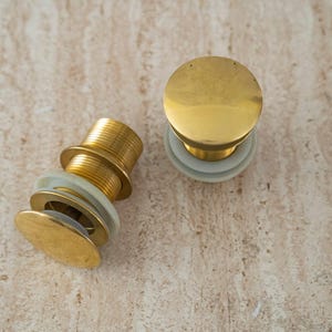 May include: Two brass sink drain stoppers with white rubber seals. One stopper is upright, and the other is on its side. The stoppers have a circular top and a threaded base. The background is a light beige, textured surface.