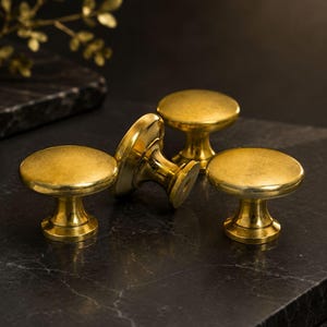 May include: A set of three round, polished brass cabinet knobs with a smooth, reflective surface. Each knob features a circular top and a tapered base, showcasing a metallic gold finish that catches the light, arranged in a cluster.