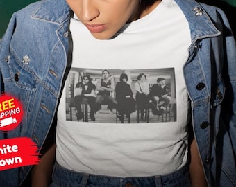 May include: A white t-shirt featuring a rectangular black and white graphic print of five individuals sitting on chairs in a row, worn under a blue denim jacket with silver buttons and a collar, showcasing a casual layered style.