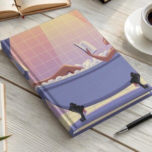 May include: A pink and purple journal with a woman reading a book in a bathtub. The journal has a white grid pattern and a blue bathtub with gold accents.