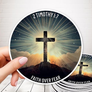 Könnte beinhalten: Ein Stapel runder Aufkleber mit einer schwarzen Silhouette eines Kreuzes auf einem Hügel mit einem Sonnenaufgang dahinter. Der Text "2 Timotheus 1:7" und "Glaube über Angst" ist auf den Aufklebern gedruckt.